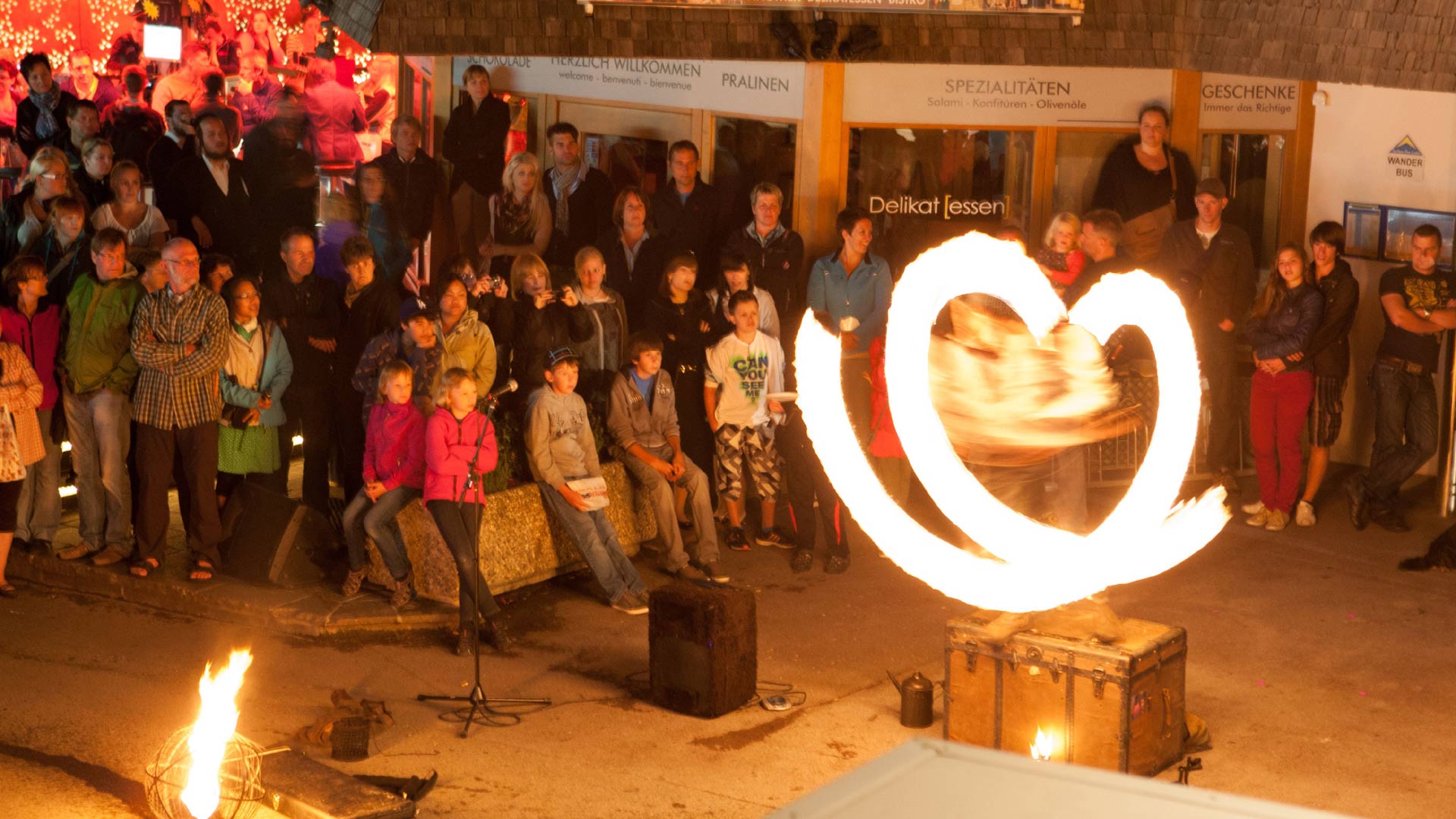 Feuershow bei Nacht mit Publikum auf Straßenfest in Serfaus, Performer formt Flammenherz.